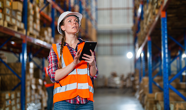 Female warehouse manager inspecting pallet racks for damage, using a tablet in a large warehouse distribution center.