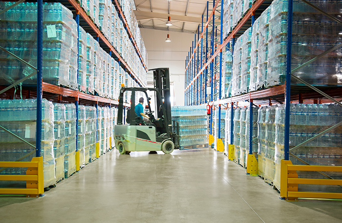forklift aisle in a warehouse, with a forklift loading pallets of water bottles.